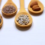 Close-up of various nuts and seeds in wooden spoons on a white background, showcasing healthy food options.