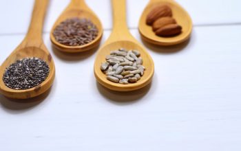 Close-up of various nuts and seeds in wooden spoons on a white background, showcasing healthy food options.