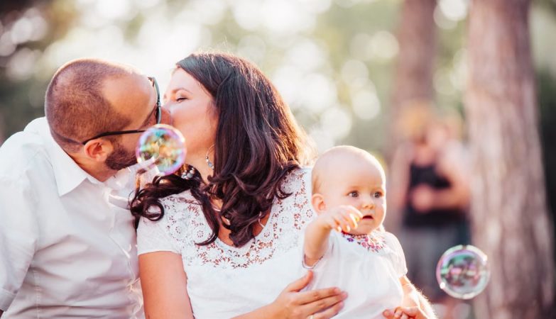 A tender family moment captured with a couple kissing and a baby holding bubbles outdoors.
