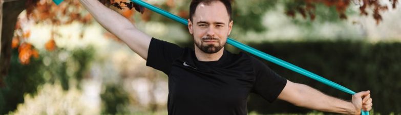 Adult man in black shirt exercises with a resistance band outside.