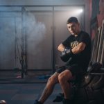 A male boxer sitting on a large tire in a gym, wearing gloves and sports attire.