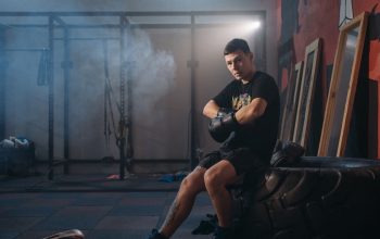 A male boxer sitting on a large tire in a gym, wearing gloves and sports attire.