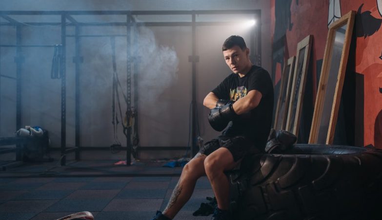 A male boxer sitting on a large tire in a gym, wearing gloves and sports attire.